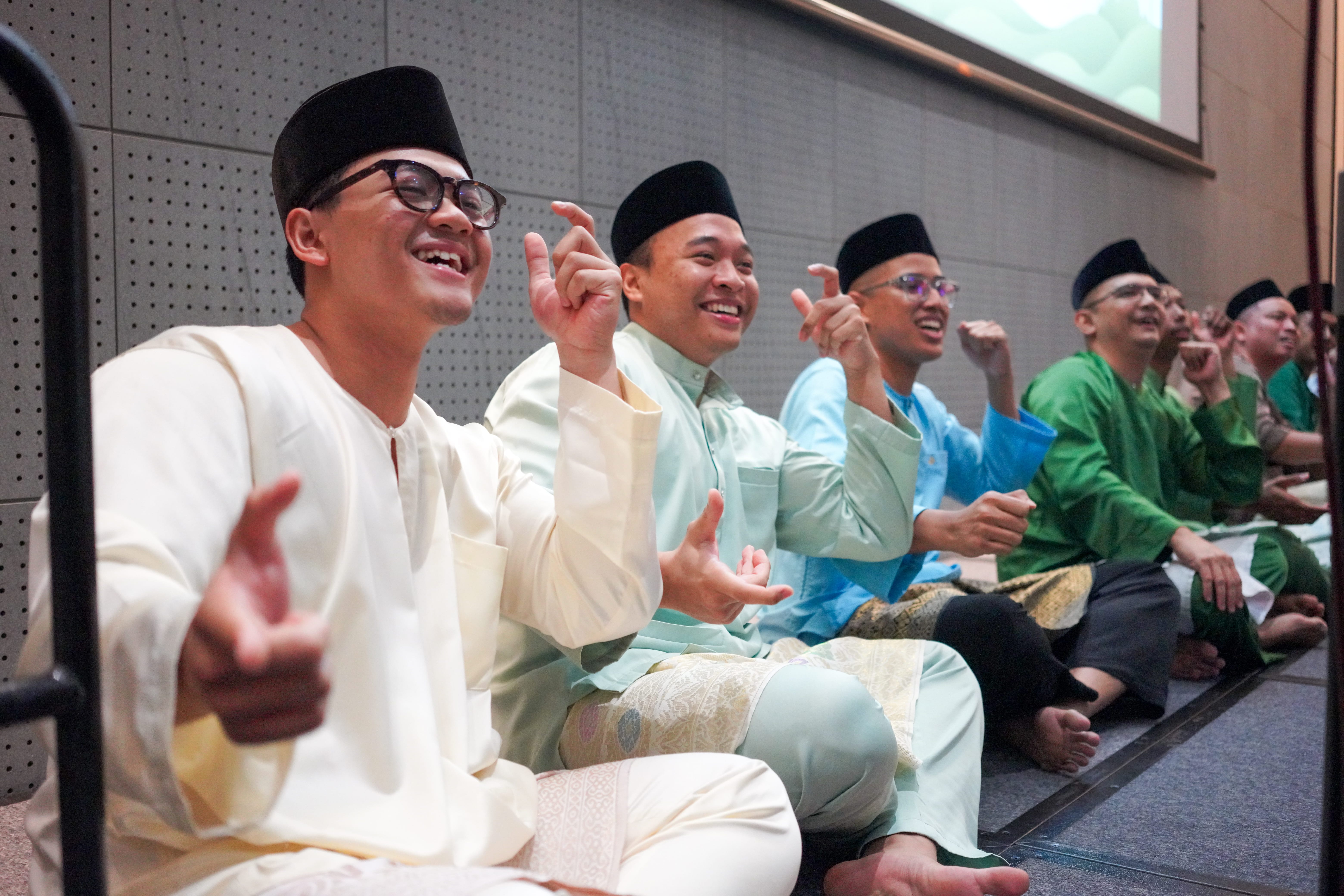 Dikir barat performers seated in a row on stage, smiling and singing while making synchronised hand gestures.